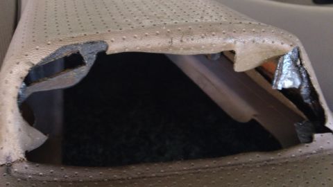 1994 Ford Bronco - Close-up of a tan perforated leather seat cushion lifted away from its frame, exposing the black metal seat pan brackets and dark carpet un…