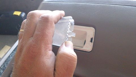 1994 Ford Bronco - A hand holds a clear textured courtesy-lamp lens next to its empty white housing recessed in a tan door panel, mid-replacement. The dashboa…