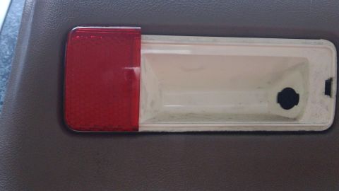 1994 Ford Bronco - A rectangular door courtesy-light housing with a red reflector lens at one end and a yellowed white lens section, removed and laid on a tan…