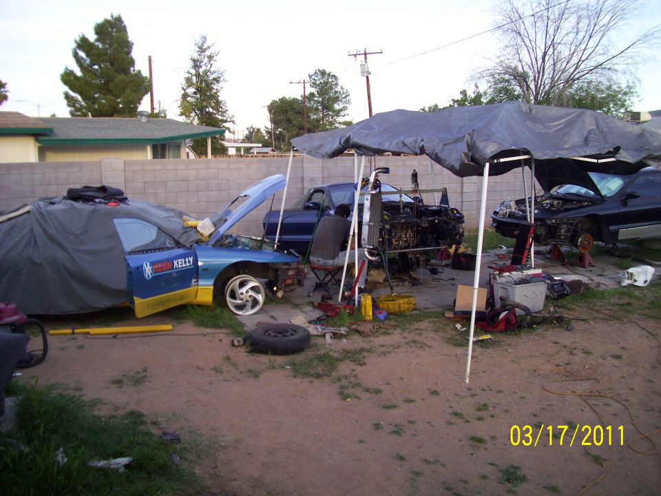 Two cars are disassembled outdoors under tarps, with parts and tools scattered around. One car has 'SPEED KELLY' on the door