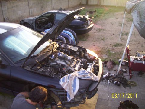 1995 Ford Probe - Two cars, one a 1995 Ford Probe, are undergoing repairs in a yard with parts and tools visible.