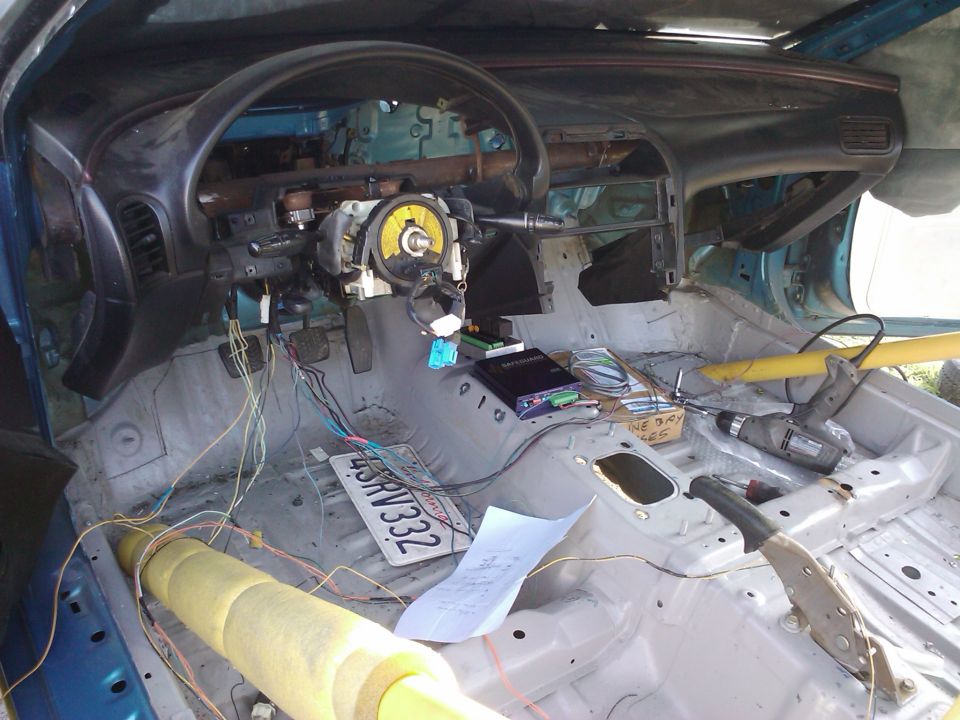 Interior of a stripped-down car with exposed wires, a drill, and a license plate on the floor