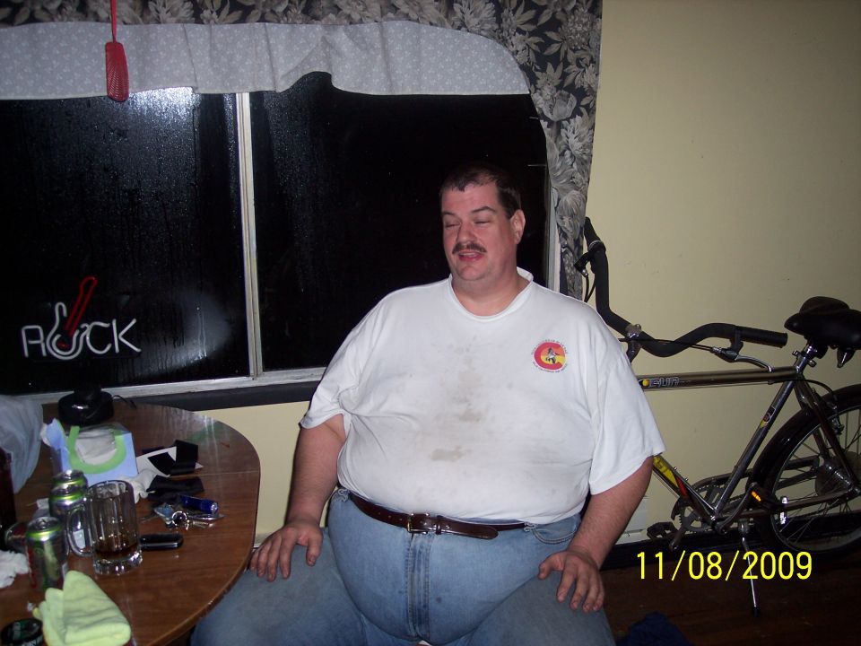 Man in white t-shirt and jeans sits at a table with drinks; bicycle behind him. Neon sign in window reads 'ROCK'