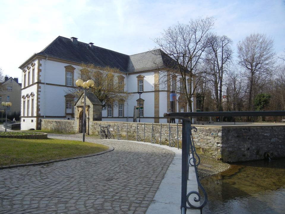 Cobblestone path leads to a large white building with a dark roof, surrounded by trees and a stone wall