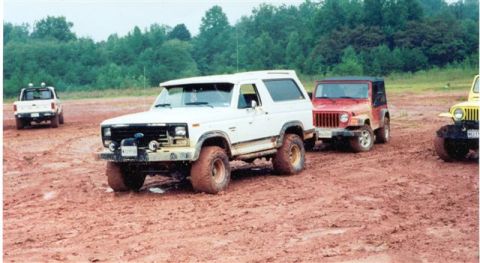 1986 Ford Bronco - Multiple off-road vehicles, including a white 1986 Ford Bronco, navigate a muddy terrain.