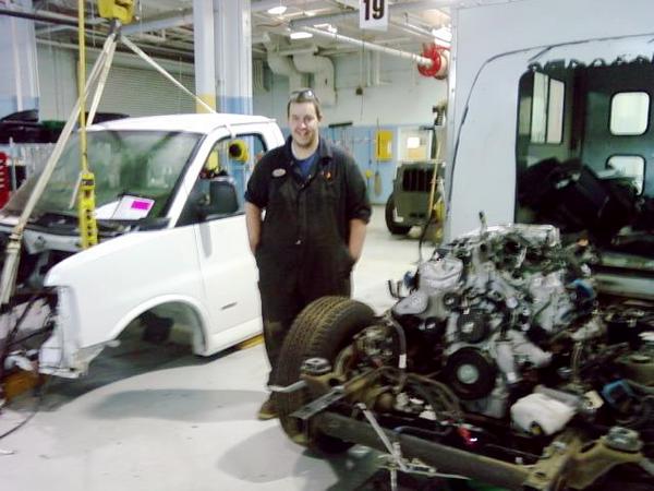 White van chassis with engine removed, a person in overalls, and a forklift in the background