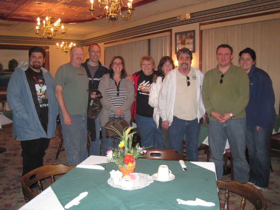 Group of nine people standing in a room with a chandelier and a table set for a meal