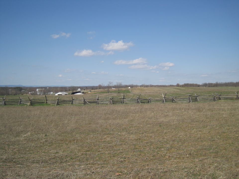 Split-rail fence in a grassy field with buildings and trees in the distance under a blue sky with clouds