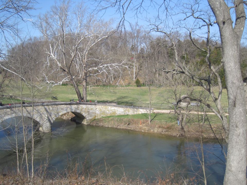 Stone arch bridge crossing a river, with bare trees and a field in the background