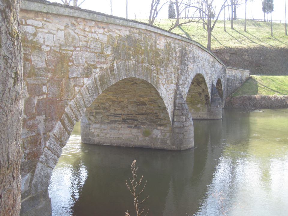 Stone arch bridge with multiple arches crossing a calm body of water, with trees and grassy hills in the background