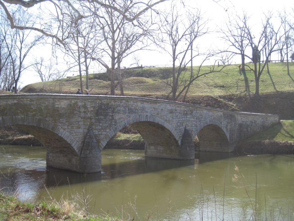 Stone arch bridge over a murky river, with bare trees and a grassy hill in the background