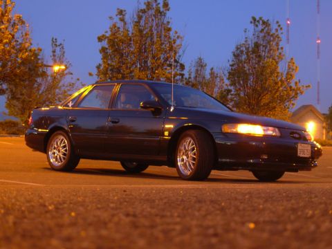 1995 Ford Taurus - A dark sedan is parked in a lot as dusk settles, with trees and streetlights in the background.