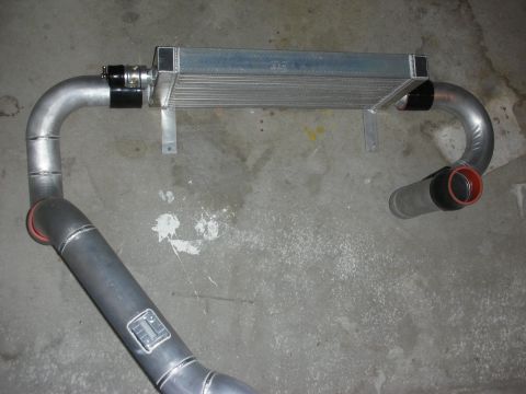 1998 Ford Crown Victoria - aluminum intercooler flanked by curved aluminum charge pipes with red-lined silicone couplers, laid on a concrete floor.