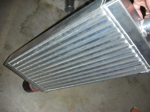 1998 Ford Crown Victoria - hand holds an aluminum bar-and-plate intercooler core with a silicone-coupled outlet, angled over a concrete floor.