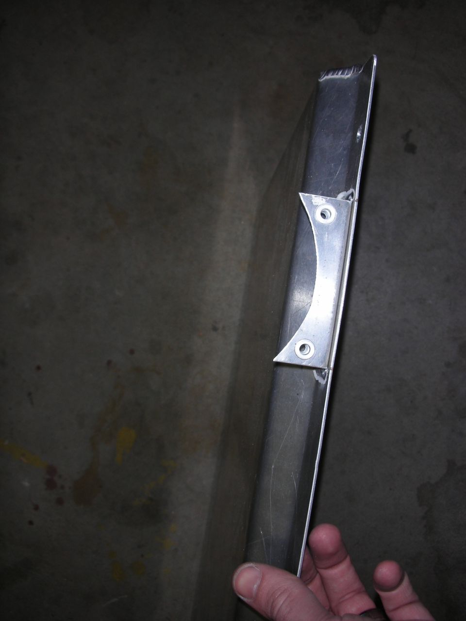 Hand holding a long aluminum extrusion with a welded bracket over concrete