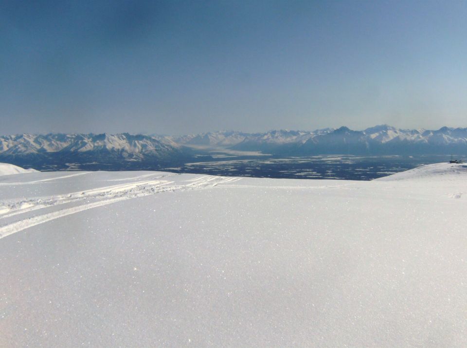 Wide view of snow-covered ground with tracks, leading to a distant valley and snow-capped mountains under a clear blue sky. Supermotors logo in corner