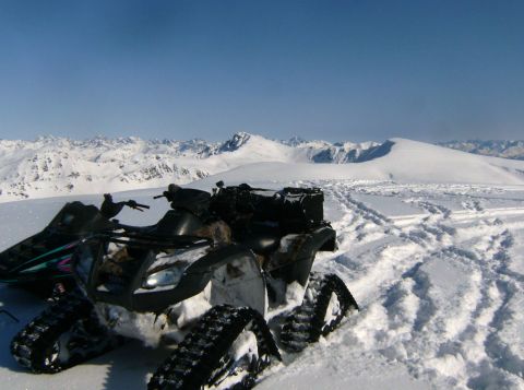 2000 Ford E350 - An ATV equipped with tracks navigates a snowy mountain landscape, with another snowmobile partially visible in the background.