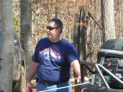 1995 Ford Bronco - A man stands by an off-road vehicle, which appears to be modified with a roll cage, in a wooded area.