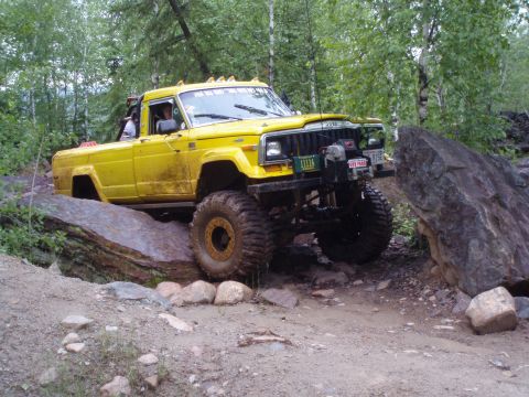 1986 Jeep J-10 - This modified Jeep J-10 is shown off-roading on a challenging, rocky path surrounded by trees.