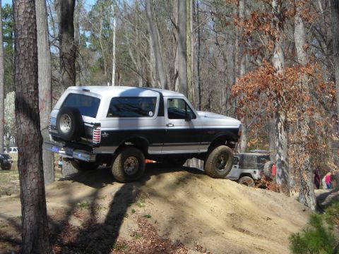 1995 Ford Bronco - A two-tone Ford Bronco navigates an off-road trail, with another vehicle and people in the background.
