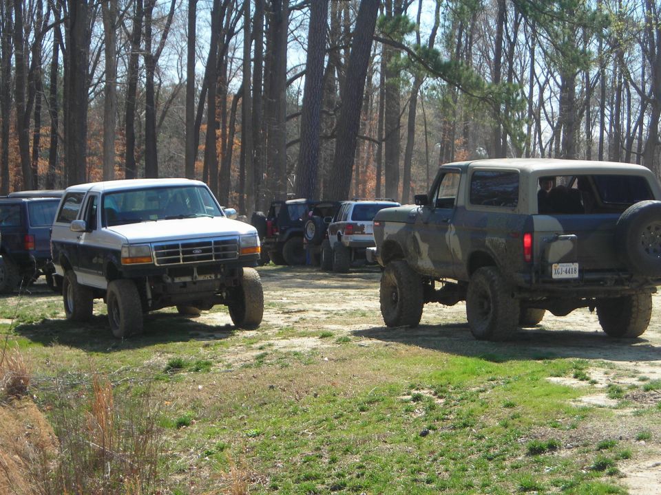 Two Ford Broncos parked in a wooded area. One is white and blue, the other is camouflaged