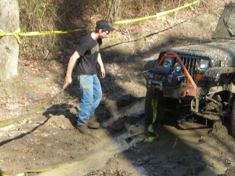 1995 Ford Bronco - A person pulls a tow strap attached to a mud-covered Jeep Wrangler stuck in a muddy off-road course.