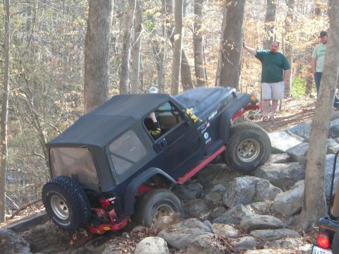 1995 Ford Bronco - A black Jeep Wrangler is shown off-roading on a challenging rocky terrain with trees in the background.