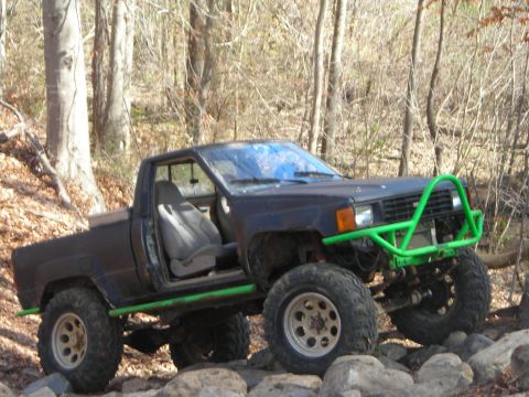 1995 Ford Bronco - This modified black pickup truck features a bright green off-road bumper and roll cage, navigating a rocky, wooded trail.