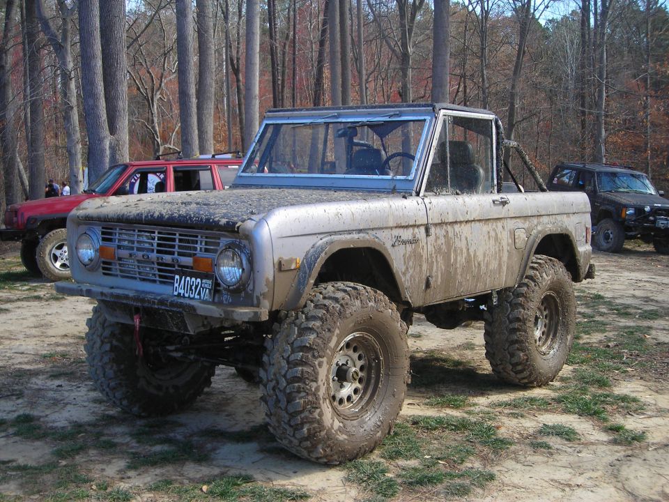 Muddy old Ford Bronco with big tires, parked with other SUVs in a wooded area