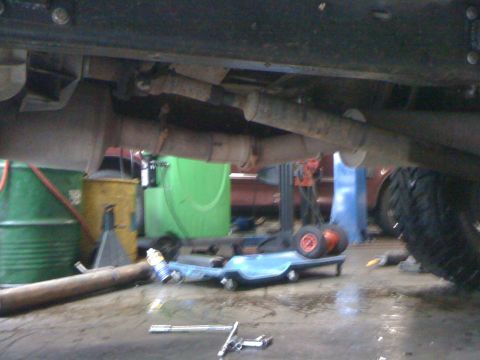 1996 Ford C-350 (Centurion Conversions) - Ground-level view under a raised truck chassis in a shop, showing the frame rails, driveshaft, muffler and exhaust piping overhead with a l…
