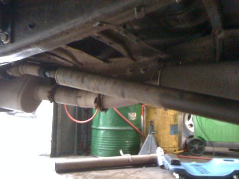 1996 Ford C-350 (Centurion Conversions) - The undercarriage of a vehicle, showing the exhaust system and drive shaft, with storage barrels visible behind.