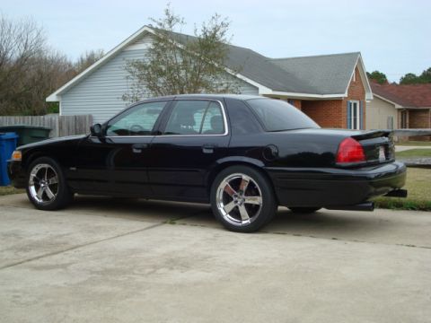 2004 Ford Crown Victoria - A black sedan with aftermarket wheels is parked in front of a house.