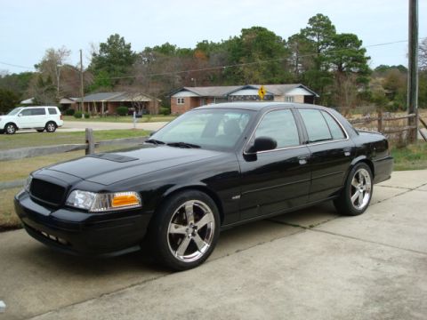 2004 Ford Crown Victoria - A black sedan with aftermarket wheels sits in a driveway, with a white SUV parked in the background.