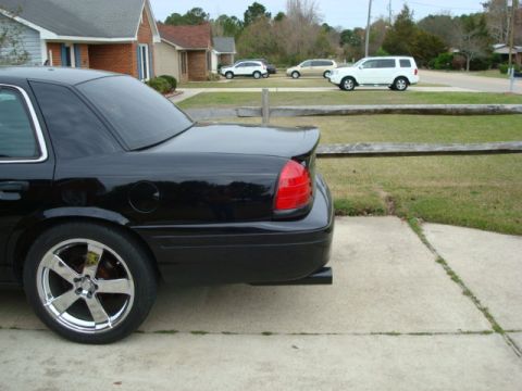 2004 Ford Crown Victoria - A black sedan with custom wheels and a spoiler is parked in a driveway, with other vehicles visible in the background.