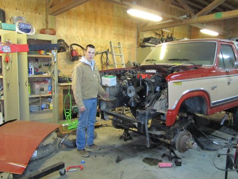 1984 Ford Bronco - Man stands beside a disassembled Ford truck in a garage, showing the vehicle's frame and removed engine.