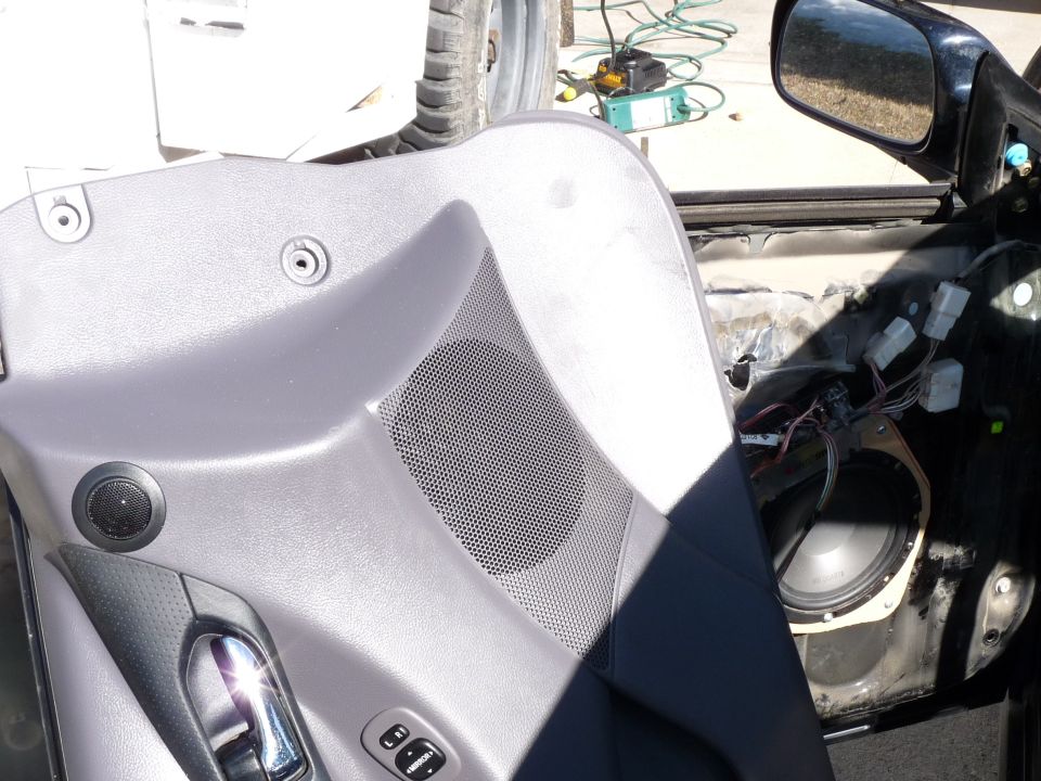 Gray car door panel with speaker grille, chrome handle, and window controls removed, revealing wiring and a speaker