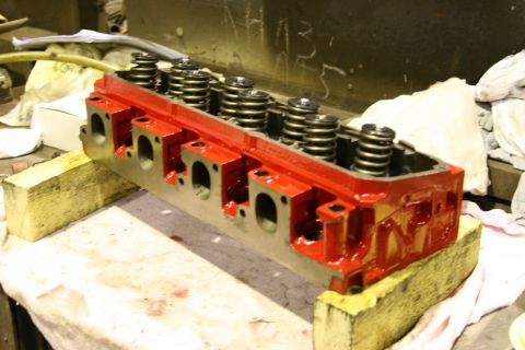 1973 Ford Ranchero - A red engine cylinder head with visible valve springs sits on wooden blocks in a workshop.