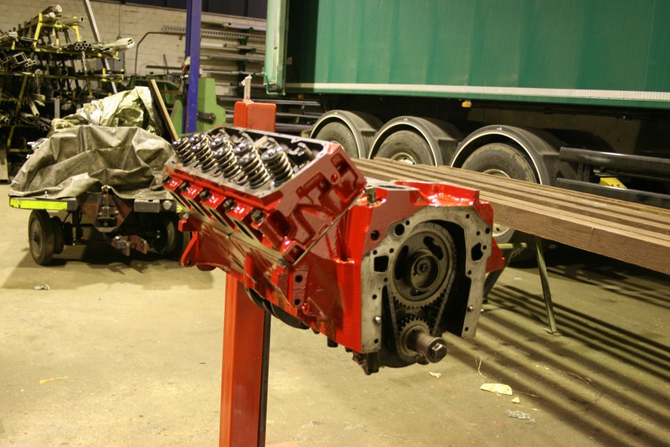 Red V8 engine block on an orange stand in a workshop, with a green truck and trailer in the background