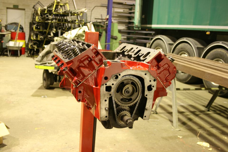 Red V8 engine block with timing gears visible, on a stand in a workshop