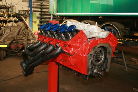 1973 Ford Ranchero - A V8 engine is displayed on a stand in a workshop setting, with a large green truck in the background.