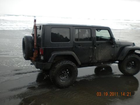 2005 Jeep Wrangler - A black Jeep Wrangler sits on a sandy beach with waves in the background, a spare tire and jack on the rear.