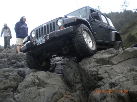 2005 Jeep Wrangler - A black Jeep Wrangler is shown off-roading on a rocky coast, with two people standing nearby.