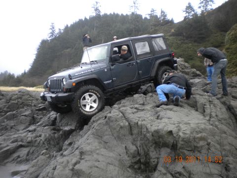 2005 Jeep Wrangler - A dark blue Jeep Wrangler Rubicon navigates rocky coastal terrain, with people observing.