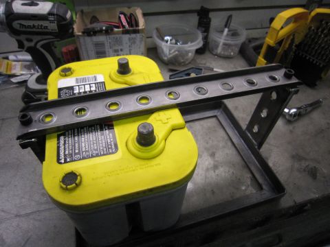 2008 Jeep Wrangler - A yellow car battery is secured by a custom metal bracket, with tools visible in the background.
