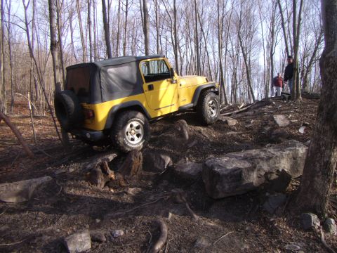 2003 Jeep Wrangler - A yellow 2003 Jeep Wrangler navigates a challenging rocky terrain in a wooded area.
