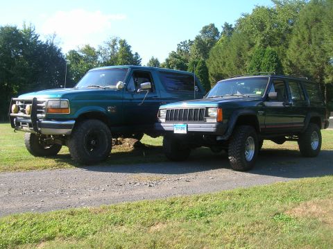 1995 Jeep Cherokee - Two SUVs, a Ford Bronco and a Jeep Cherokee, are parked on a gravel driveway with trees in the background.