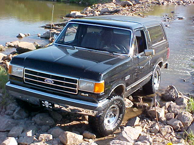 Black Ford Bronco with a skull and crossbones license plate, driving over rocks in a stream
