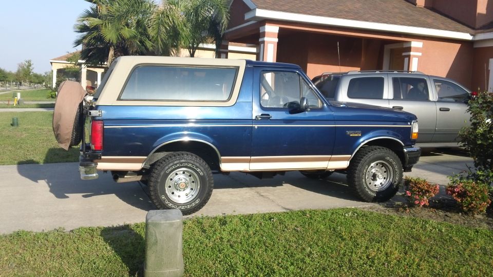 1994 Ford Bronco, blue with tan removable top, spare tire mounted on rear door. Silver SUV parked behind