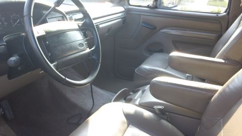 1994 Ford Bronco - Interior shot from the driver's side of a 1990s Ford truck showing the black four-spoke airbag steering wheel, tan dash pad and door panel,…