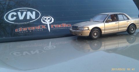 2000 Mercury Grand Marquis - A small toy car sits on a reflective surface with several stickers on a dark background.